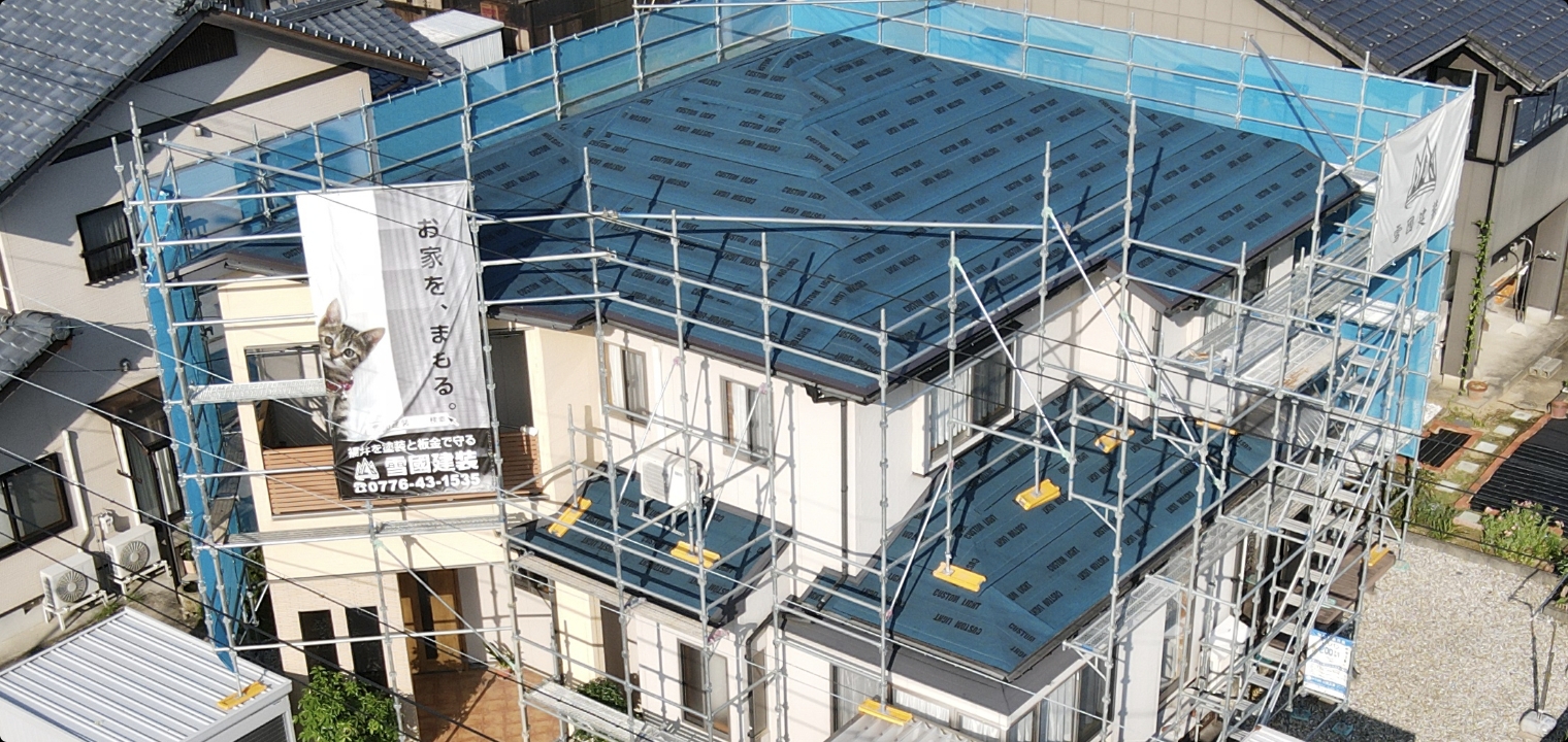 住宅の屋根と外壁を囲む足場と、雪國建装の現場幕